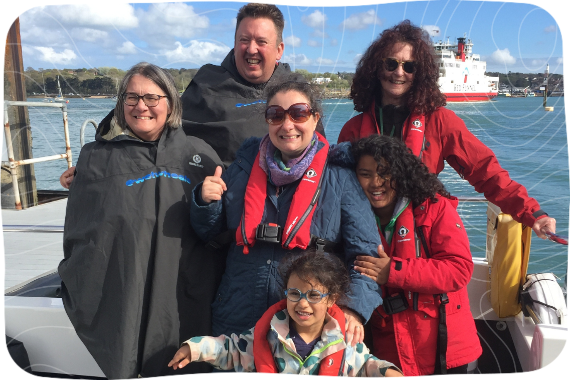 A photograph of Angela, Esther, their children, and Dementia Adventure volunteers on holiday together. They're standing on a boat with a view of the water behind them.
