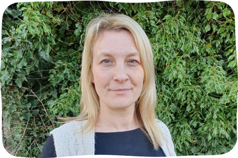 Karen, a blonde Caucasian woman standing in front of a green bush.