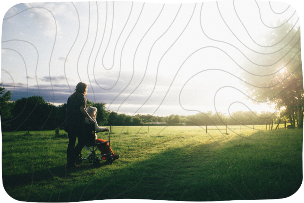 Two women silhouetted, one pushing the other in a wheelchair, staring out at a sunrise over green fields.